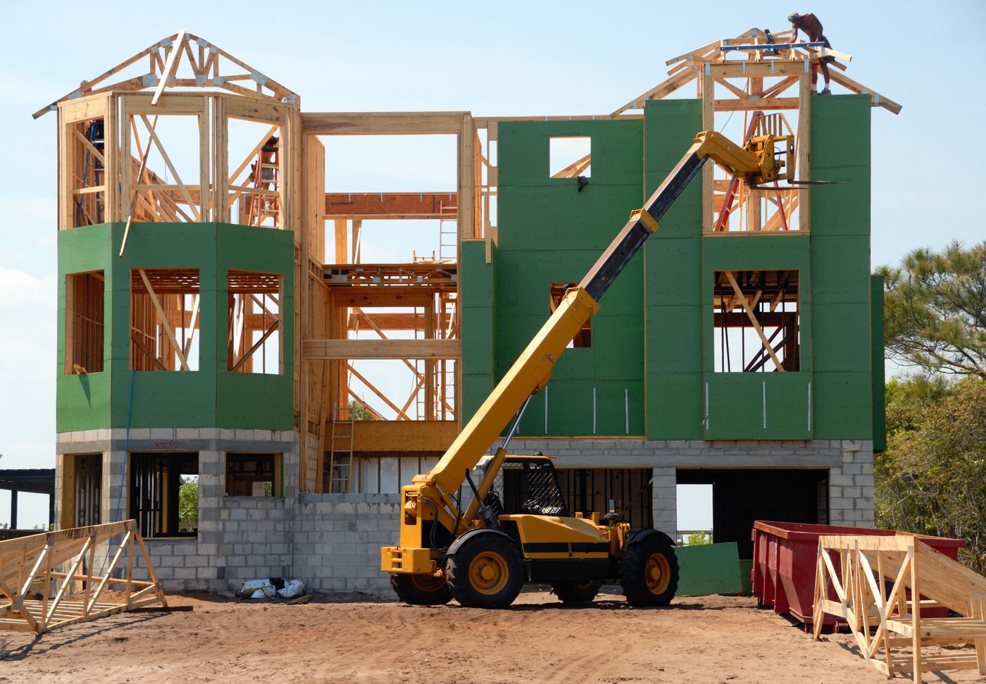 Residential build under construction with yellow telehandler raised toward exposed roof trusses and a worker on the peak
